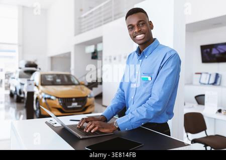 Venditore di auto Smiling Black che utilizza un computer portatile seduto a un tavolo con un tablet nello showroom Foto Stock