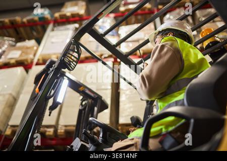 Lavoratrice mista che indossa attrezzature di sicurezza utilizzando la radio durante il funzionamento del carrello elevatore a forche in magazzino Foto Stock