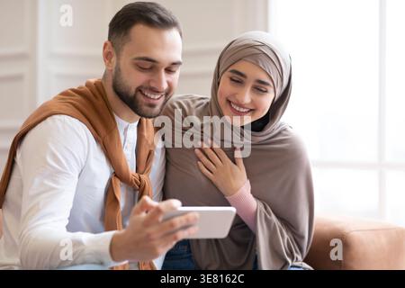 Donna musulmana araba in hijab e marito caucasico che guardano divertenti video sullo smartphone mentre si siede insieme sul divano nel soggiorno di casa Foto Stock