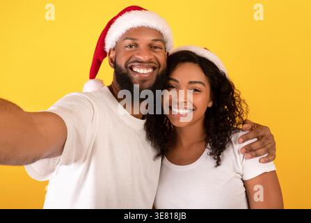 Coppia nera sorridente che indossa i cappelli di Babbo Natale scattando selfie su sfondo giallo in studio Foto Stock