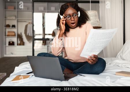 Donna nera scioccata con capelli ricci che indossano cuffie e occhiali che tengono e guardano la carta mentre si siede sul letto con un computer portatile in camera da letto a casa Foto Stock