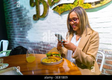 Donna d'affari ispanica sorridente che indossa gli occhiali da vista con lo smartphone mentre siede al tavolo con un hamburger al ristorante Foto Stock