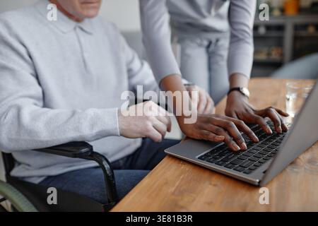 Immagine ritagliata di un'infermiera nera che aiuta un paziente di mezza età caucasico a digitare sul laptop accanto a lui a casa Foto Stock