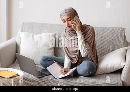 Donna musulmana araba in hijab che scrive appunti mentre fa una telefonata seduto con le gambe incrociate sul divano con un portatile a casa Foto Stock