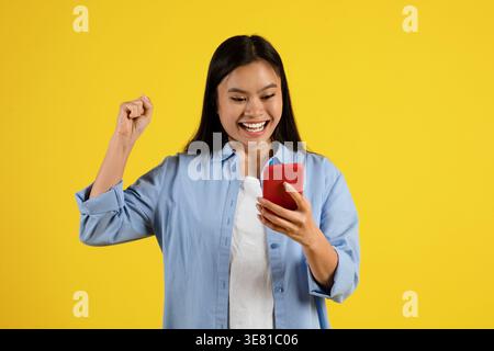 Adolescente asiatica sorridente con pugno stringato che utilizza lo smartphone mentre si trova sullo sfondo giallo in studio Foto Stock