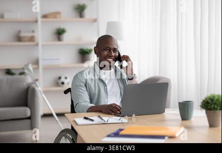 Allegro disabile uomo d'affari nero calvo in sedia a rotelle che ha una telefonata al tavolo con un computer portatile e documenti nel soggiorno di casa Foto Stock