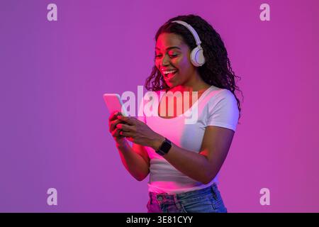 Donna nera che indossa le cuffie con smartphone mentre si trova sullo sfondo viola e rosa in studio Foto Stock