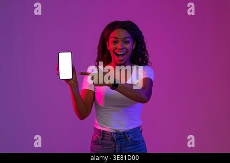 Donna nera eccitata con camicia bianca e jeans che tengono e puntano sullo schermo vuoto dello smartphone mentre si sta in piedi su sfondo viola e rosa io... Foto Stock
