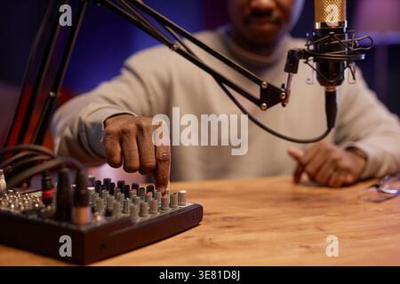 Immagine ritagliata dei controlli di commutazione del podcaster maschio nero sulla scheda del mixer audio mentre si è seduti alla scrivania con il microfono in studio Foto Stock