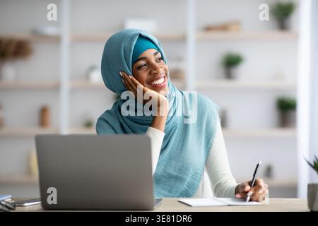Una ragazza nera sorridente che indossa un hijab blu che guarda lontano e scrive appunti mentre si siede a un tavolo con il laptop a casa Foto Stock