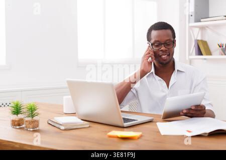 Uomo di colore che indossa gli occhiali da vista che fa una telefonata e legge i documenti seduti a un tavolo con un computer portatile e documenti in ufficio Foto Stock