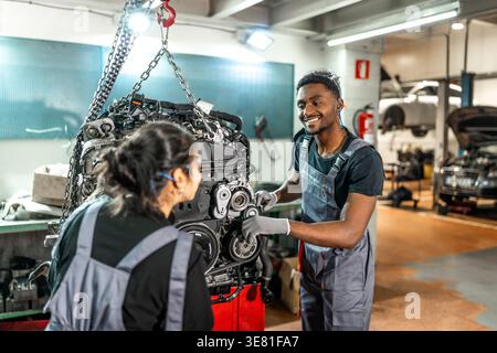 Meccanici neri maschi e asiatici che indossano uniformi e occhiali protettivi che ispezionano il motore di un'auto appeso a un paranco a catena in un negozio di riparazione auto Foto Stock