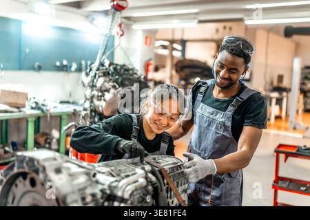 Meccanici neri maschi e asiatici che indossano uniformi e occhiali protettivi assemblando il motore di auto pLifestyles in officina di riparazione auto Foto Stock