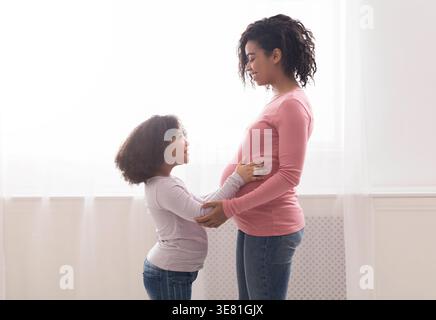 Ragazza nera che tocca la pancia di madre incinta in piedi contro la finestra a casa Foto Stock