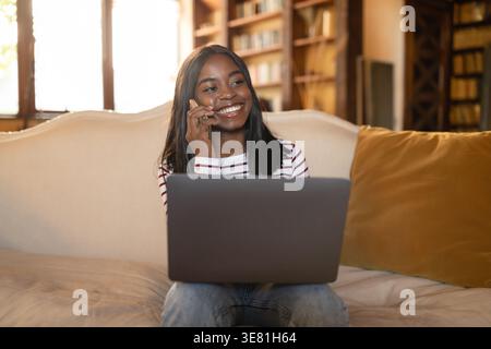 Adolescente di colore che riceve una telefonata seduto con un laptop sul divano nel soggiorno di casa Foto Stock