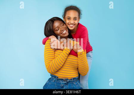 Cheerful Black pre-teen girl in piedi e abbracciare la madre da dietro su sfondo blu in studio Foto Stock
