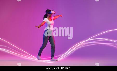 Donna nera che indossa una camicia bianca e un visore VR che sperimenta la realtà virtuale mentre cammina in linee luminose al neon sullo sfondo viola nel ... Foto Stock