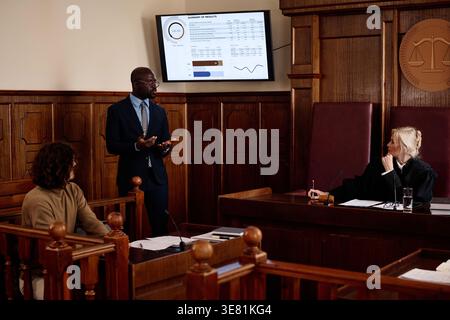 Avvocato di sesso maschile di colore in piedi e presentare il sommario dei risultati mentre spiega i dati a giudice, giuria e imputato durante la seduta del processo... Foto Stock