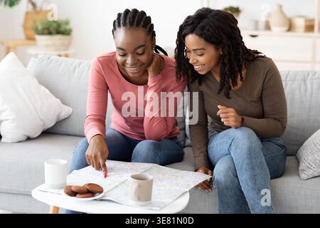 Amiche nere che guardano la mappa mentre si siedono sul divano nel soggiorno di casa Foto Stock