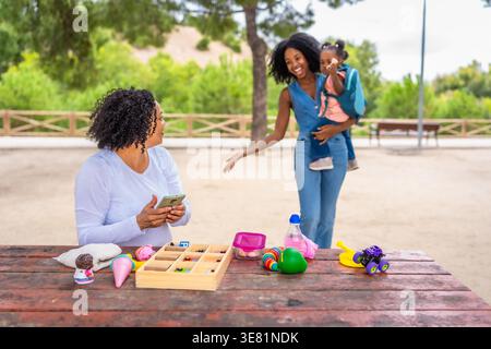 La nonna ispanica seduta a tavola guardando la figlia nera che porta la bambina che cammina verso di lei nel parco durante il giorno Foto Stock