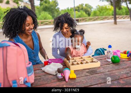 Bambina nera che gioca con i giocattoli con la madre e la nonna sedute sulla panchina nel parco durante il giorno Foto Stock