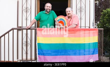 Sorridente coppia di uomini ispanici e caucasici di mezza età in piedi insieme sul balcone con bandiera arcobaleno e ventilatore a casa durante il giorno Foto Stock