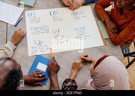 Gruppo di studenti immigrati seduti a tavola che imparano verbi frastagliati durante la lezione di inglese in biblioteca Foto Stock