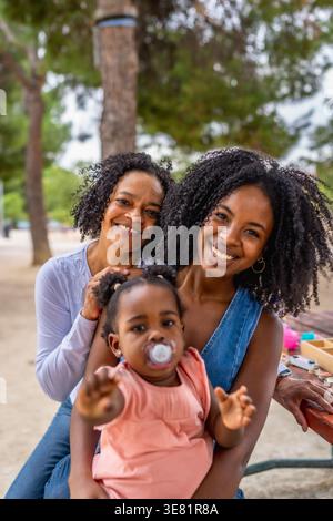 Bambina nera con succhietto seduta con mamma e nonna ispanica nel parco durante il giorno Foto Stock