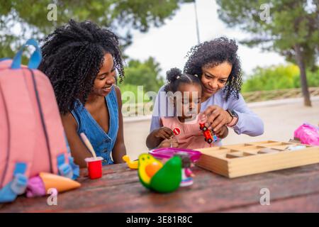 Nonna ispanica che gioca con la nipotina mentre la mamma nera li guarda seduti a tavola nel parco durante il giorno Foto Stock
