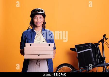 Ritratto della donna che indossa il casco e la divisa pizzeria che trasporta le scatole della pizza durante la consegna del takeaway. Dipendente asiatico del ristorante sorridente alla macchina fotografica in studio con sfondo giallo Foto Stock