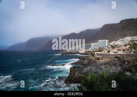 Un arcobaleno si estende sull’Oceano Atlantico davanti alle scogliere di Acantilados de los Gigantes a Puerto de Santiago, comune di Santiago del Teide, sulla costa sud-ovest di Tenerife nelle Isole Canarie, Spagna. Hotel e appartamenti bianchi costeggiano la riva vulcanica mentre la nebbia si sposta lungo la scogliera di basalto dopo una pioggia invernale. Foto Stock
