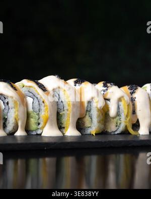 Panini di sushi glisteno con salsa cremosa e sesamo nero. Ogni rotolo presenta riso, alghe e vivaci ripieni gialli. Lo sfondo scuro evidenzia Foto Stock