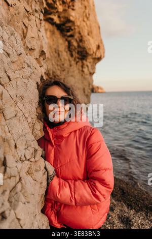 Donna vacanza costiera giovane donna in giacca rosa con pompino e occhiali da sole appoggiati sulla scogliera rocciosa vicino al mare calmo durante il tramonto Foto Stock
