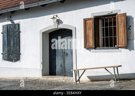 Un edificio rustico con una grande porta di metallo, persiane intemprate e una finestra sbarrata. Una semplice panca di legno siede all'esterno, invitante riposo. La scena evoca Foto Stock