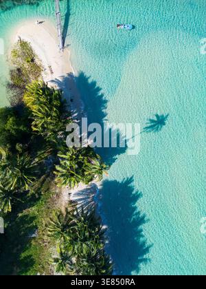 Vista aerea con droni della spiaggia tropicale dell'isola con palme, laguna turchese e molo di legno che si estende in acque cristalline e poco profonde, i bungalow del resort si nascondono Foto Stock