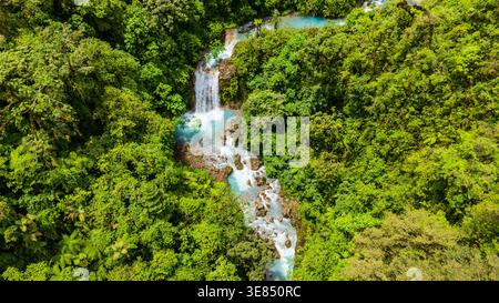 Vista aerea della lussureggiante foresta pluviale tropicale con rapide turchesi del fiume e un piccolo ponte di legno che attraversa un ruscello roccioso. Foto Stock