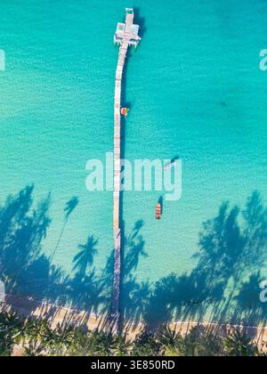 Vista aerea dall'alto verso il basso di un lungo molo che si estende nel mare turchese con piccole barche nelle vicinanze e ombre di palme lungo una costa tropicale sabbiosa. Foto Stock