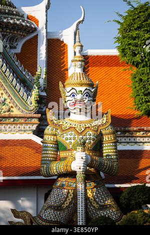 Statua decorata del guardiano presso il tempio buddista tailandese, piastrelle a mosaico e dettagli in oro, personale di supporto, tetto piastrellato rosso e cielo blu chiaro, architetto tradizionale Foto Stock