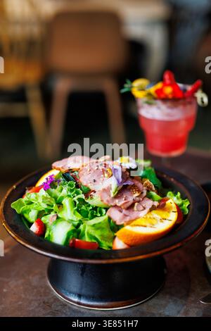 Insalata gourmet con arrosto a fette, verdure, frutta e noci in una ciotola nera; sfondo caffè sfocato con un cocktail di frutta rossa e paglia. Foto Stock