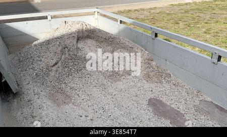 Un mucchio di ghiaia e sabbia nella parte posteriore del pianale di un camion in un cantiere con un'area erbosa e una strada visibile sullo sfondo Foto Stock