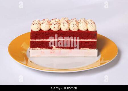 Una fetta di torta di velluto rosso condita con spugne panna montata e spruzzi rossi, servita su un piatto giallo e bianco su una tovaglia bianca. Foto Stock
