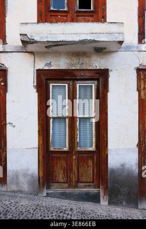 Primo piano di un vecchio ingresso di un edificio con una porta di legno intemprata, pannelli di vetro e vernice sfaldante su una parete di stucco incrinata, che mostra il decadimento urbano e vi Foto Stock