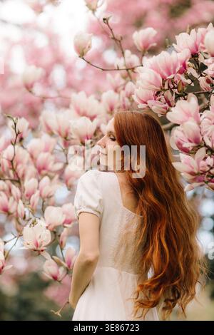 Ritratto di una giovane donna dai capelli rossi in un abito bianco in piedi tra rami di magnolia rosa in fiore in primavera, morbido sfondo bokeh, sereno romantico Foto Stock