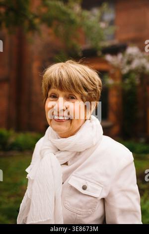 Caldo ritratto di una donna anziana sorridente che indossa una sciarpa bianca e una giacca leggera all'aperto, con un morbido verde bokeh e un'architettura in mattoni sullo schienale Foto Stock