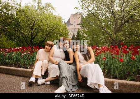 Quattro donne di età diverse siedono insieme accanto a un letto di tulipani rossi in un parco cittadino, sorridenti e appoggiate l'una sull'altra, con alberi lussureggianti e un bui storico Foto Stock
