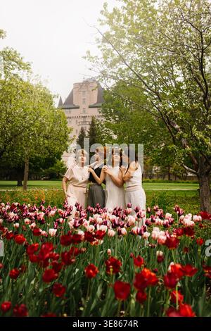 Quattro donne di età diverse condividono un caldo abbraccio in un lussureggiante parco primaverile, in piedi con tulipani rossi e rosa fioriti con alberi verdi e un Grand hotel i Foto Stock