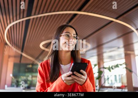 Giovane donna sicura di sé con un blazer arancione brillante che regge uno smartphone, in un ambiente aziendale moderno. Trasuda positività e prontezza per la p Foto Stock