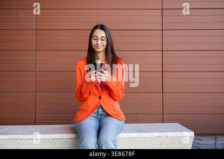 Donna d'affari con un blazer arancione seduto su una panchina, concentrata sul suo smartphone. Sorride dolcemente mentre interagisce con il suo dispositivo mobile, contro un m Foto Stock