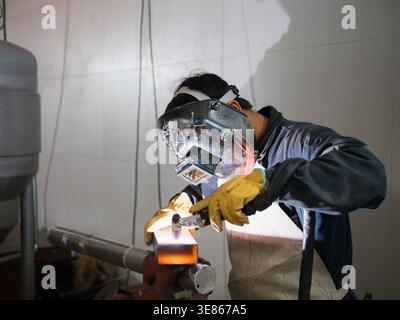 L'uomo che lavora come saldatore utilizza casco e guanti protettivi mentre unisce parti metalliche con torcia in un'officina industriale, concentrandosi sulla sicurezza e sul precis Foto Stock
