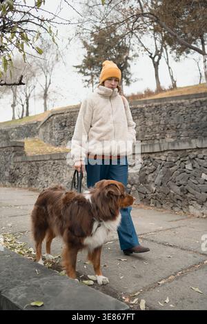 Donna in giacca calda e berretto che cammina un pastore australiano al guinzaglio lungo un tranquillo marciapiede urbano accanto a un muro di pietra, catturando la calma piaga autunnale Foto Stock